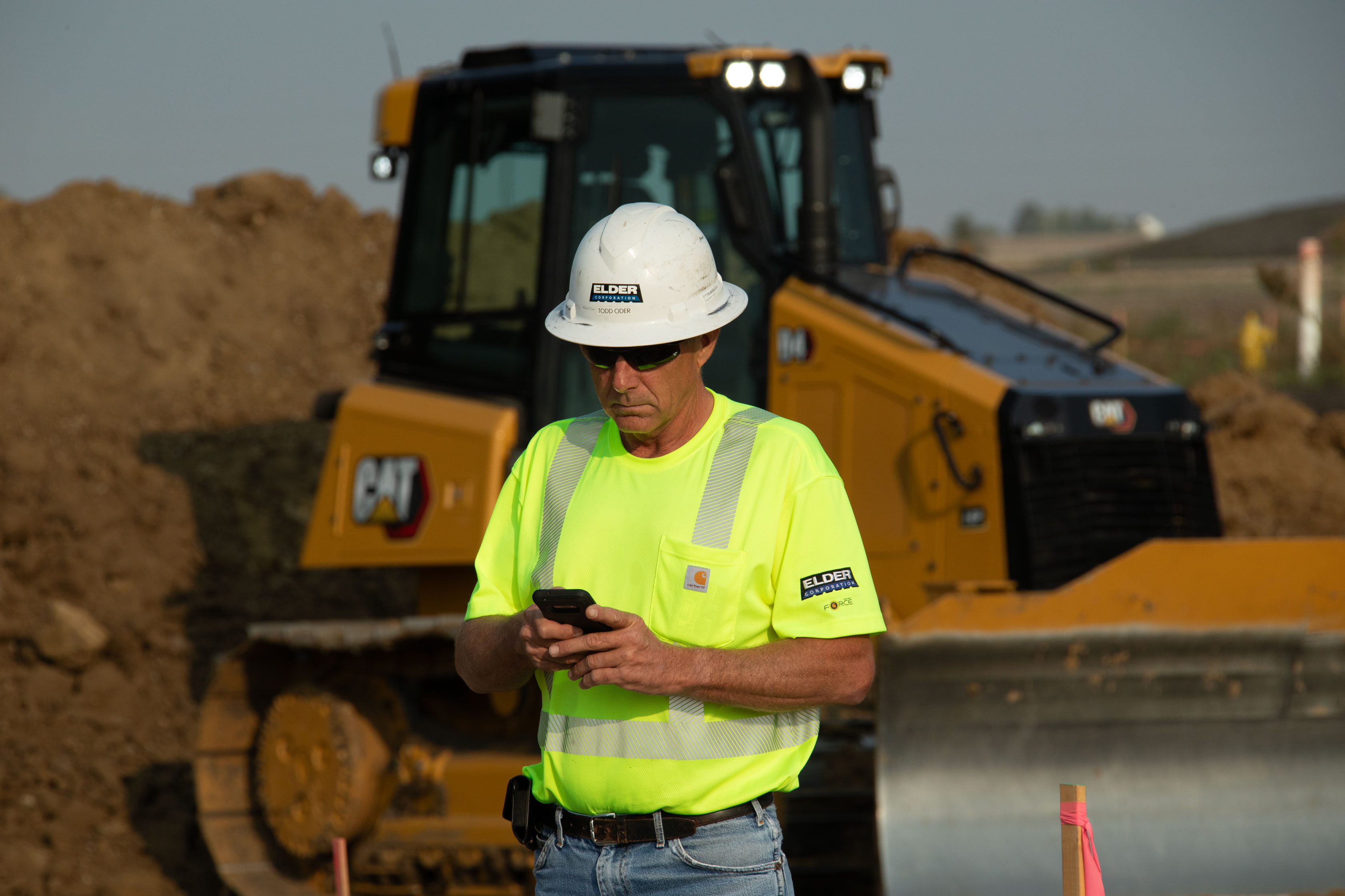A construction worker looking at a phone with construction equipment in the background.