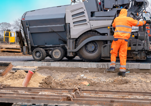 Asphalt paver filled with hot tarmac laying new road surface on new residential housing development site and roadworker operator in orange hi-viz next to it