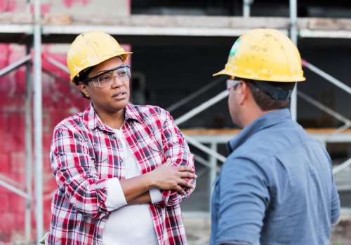 Two multi-ethnic construction workers meeting at a construction site, talking about a serious matter. One of them, a mature African-American woman in her 40s is standing with her arms crossed, looking at the other one, a young Hispanic man.