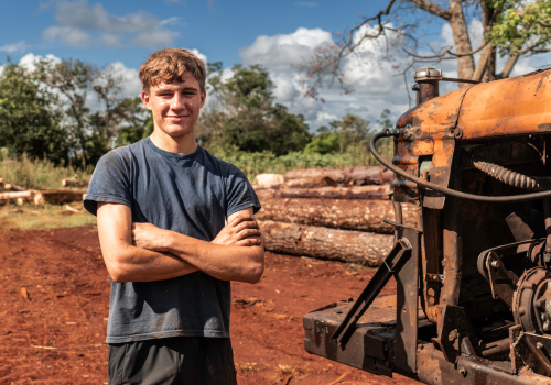 Portrait of young man working in a sawmill smiling and looking at the camera in a big field with a tractor next to him.